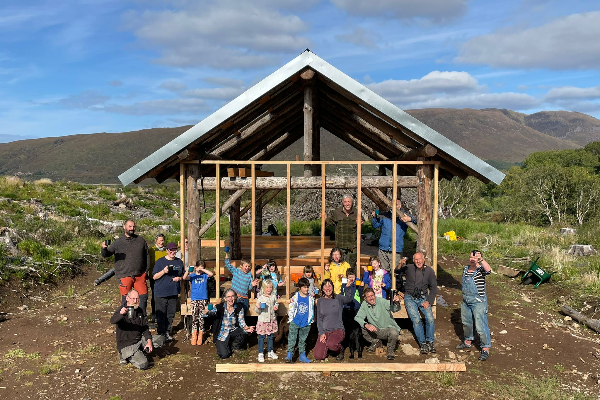 Community Woodland volunteers sitting in front of the shelter in Torgarve on a sunny day, holding up cups